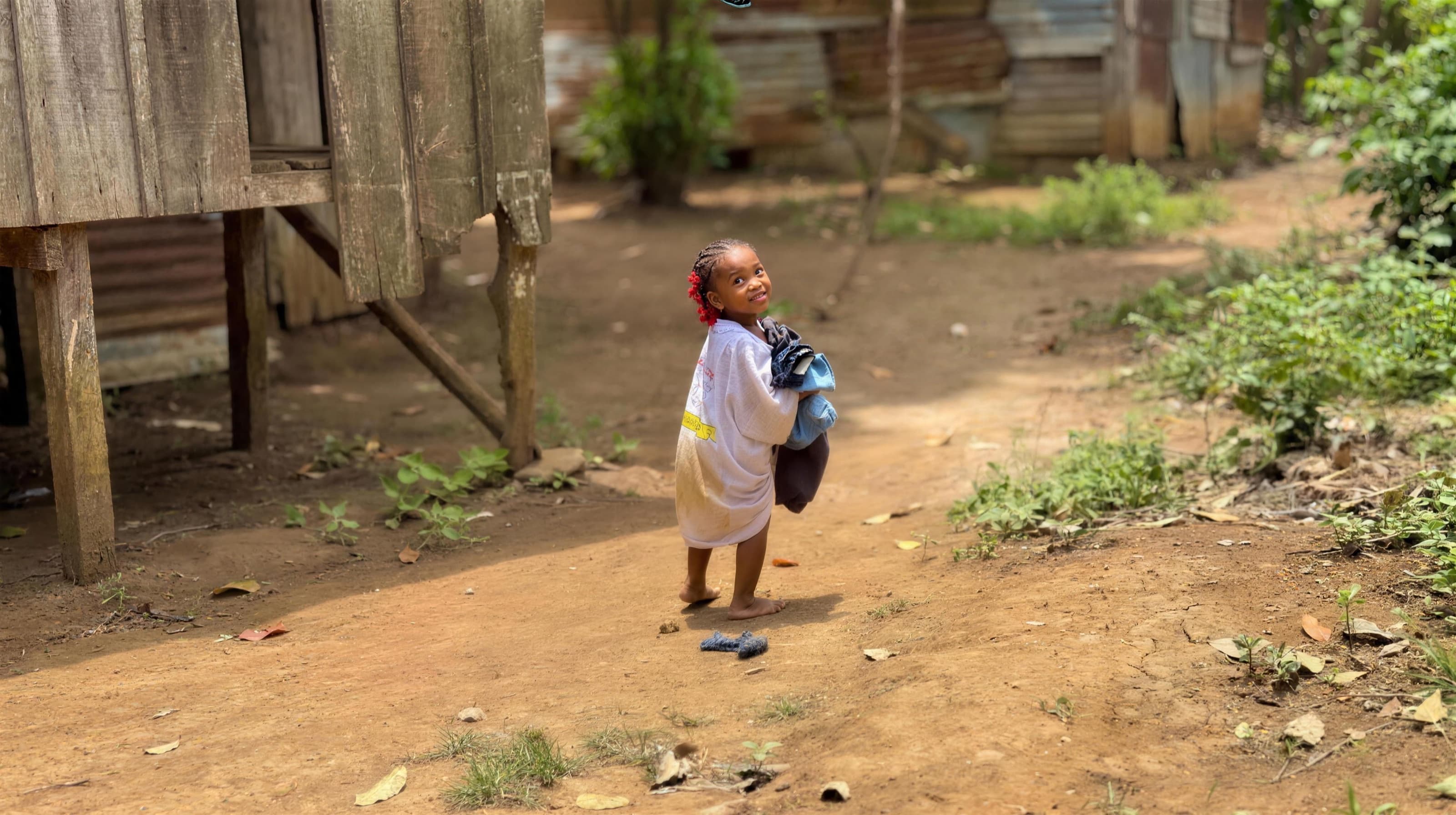 Uma menina descalça num caminho de terra numa aldeia de São Tomé, a segurar uma trouxa de roupa e a olhar para cima com um sorriso.